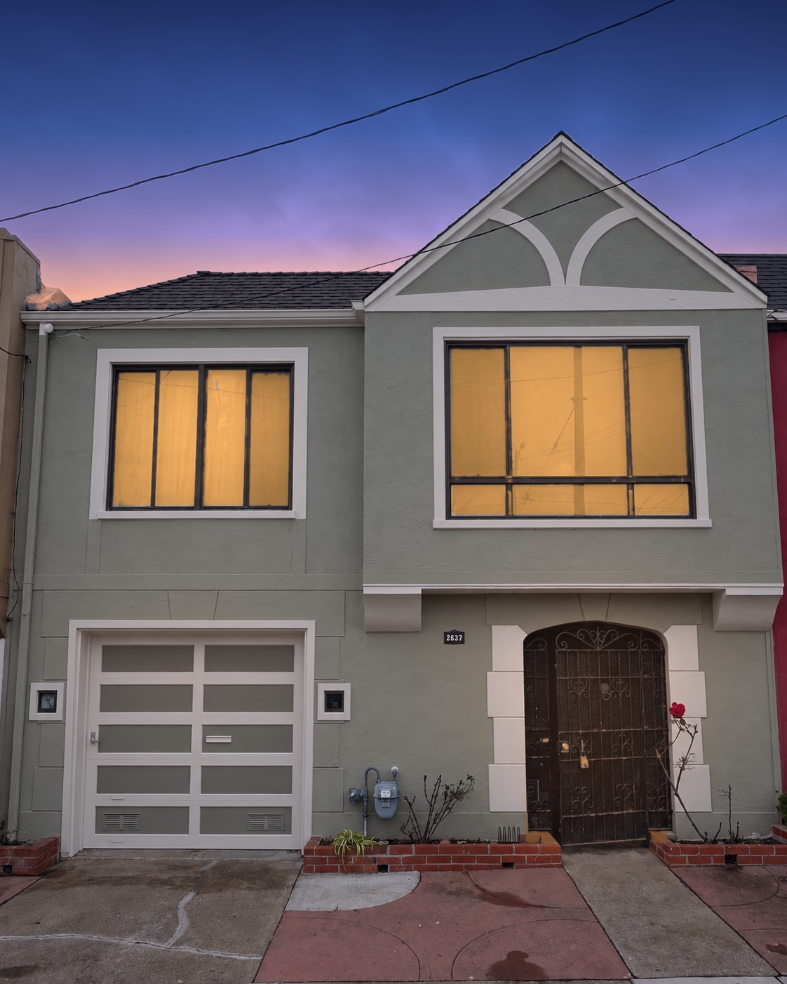 Front of the home at dusk (virtually enhanced twilight): a two-story sage-green stucco house with cream trim, with a gabled bay window, single-car garage, and an arched wrought-iron entry gate